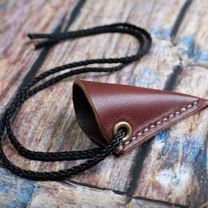 May include: A brown leather knife sheath with a black cord. The sheath has a pointed tip and a small brass eyelet.