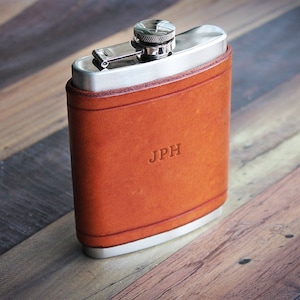 May include: A hip flask with a brown leather casing and a stainless steel top. The initials "JPH" are embossed on the leather. The flask is presented on a wooden surface.