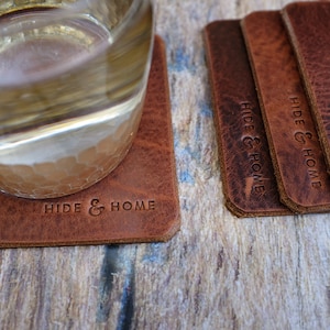May include: A set of rectangular brown leather coasters with the words "HIDE & HOME" embossed on them. One coaster has a glass on it. The coasters are on a wooden surface.