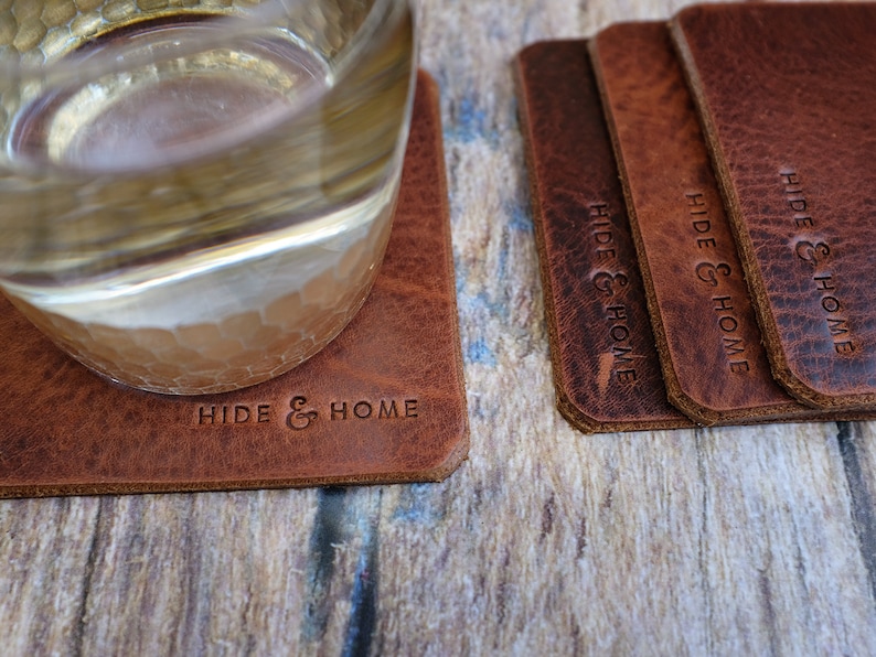 May include: A set of rectangular brown leather coasters with the words "HIDE & HOME" embossed on them. One coaster has a glass on it. The coasters are on a wooden surface.