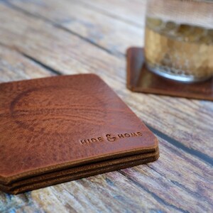 May include: A stack of square, brown leather coasters with the embossed text "HIDE & HOME". The coasters have a textured surface and are stacked on a wooden surface. A glass of liquid sits on a matching coaster.