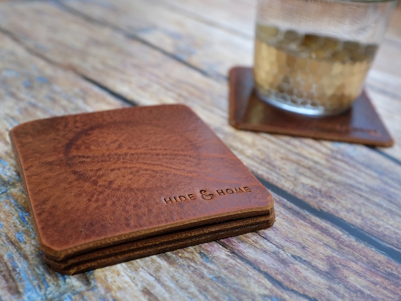 May include: A stack of square, brown leather coasters with the embossed text "HIDE & HOME". The coasters have a textured surface and are stacked on a wooden surface. A glass of liquid sits on a matching coaster.