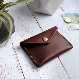 May include: A brown leather wallet with a flap closure and a brass button. The wallet is on a white wooden surface.