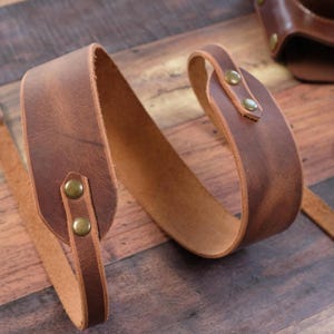 May include: A brown leather camera strap with brass accents and a ring on one end. The strap is folded and laid on a wooden surface.