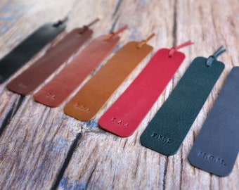 May include: Seven leather bookmarks in different colors with personalized initials. The bookmarks are arranged in a row from left to right, with the black bookmark on the left and the dark blue bookmark on the right. The initials are embossed on the bookmarks.