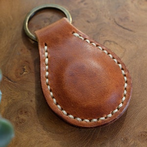 Peut inclure: Un porte-clés en cuir marron avec une bordure cousue blanche. Le porte-clés a un anneau en métal.