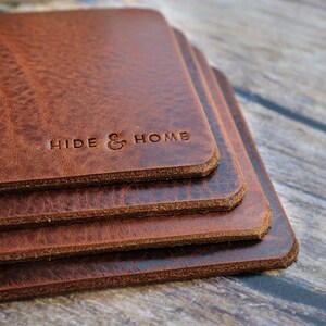 May include: A stack of four square, brown leather coasters. The coasters have a textured surface and rounded edges. The words "HIDE & HOME" are embossed on the top coaster. The coasters are stacked on top of each other.