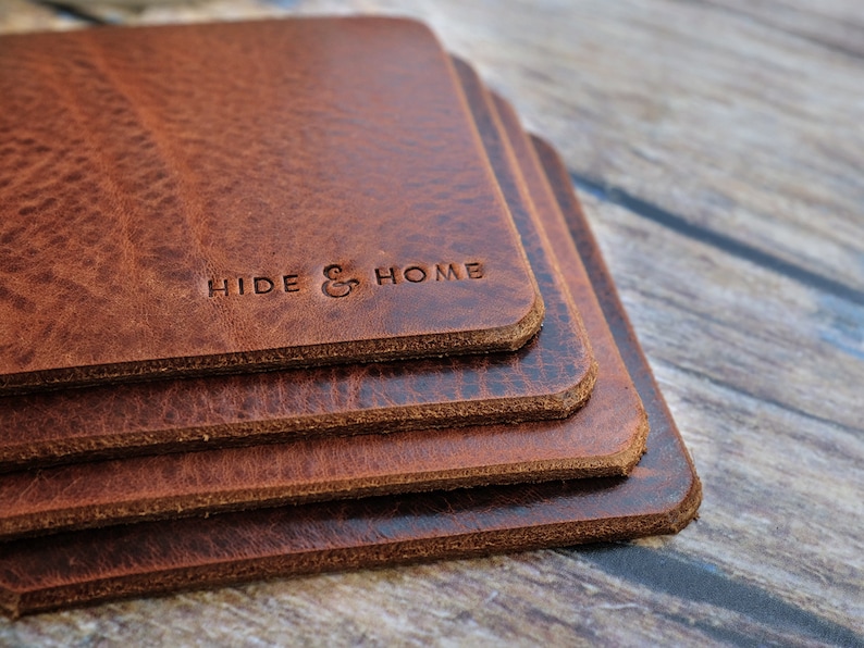 May include: A stack of four square, brown leather coasters. The coasters have a textured surface and rounded edges. The words "HIDE & HOME" are embossed on the top coaster. The coasters are stacked on top of each other.