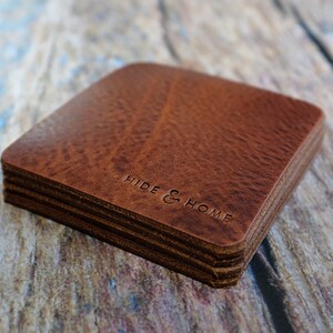 May include: A stack of square, brown leather coasters with rounded corners. The coasters have a textured surface and the words "HIDE & HOME" are embossed on the top coaster. The coasters are stacked on a wooden surface.