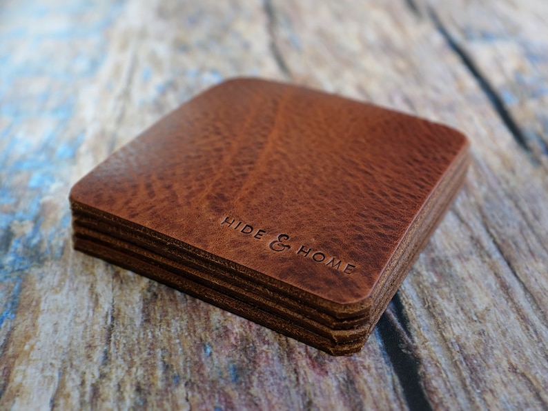 May include: A stack of square, brown leather coasters with rounded corners. The coasters have a textured surface and the words "HIDE & HOME" are embossed on the top coaster. The coasters are stacked on a wooden surface.