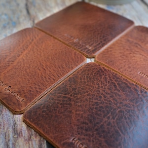 May include: Four square brown leather coasters arranged in a flower-like pattern. The coasters have a textured surface and the words "HIDE & HOME" are embossed on the surface. The coasters are a warm brown color.
