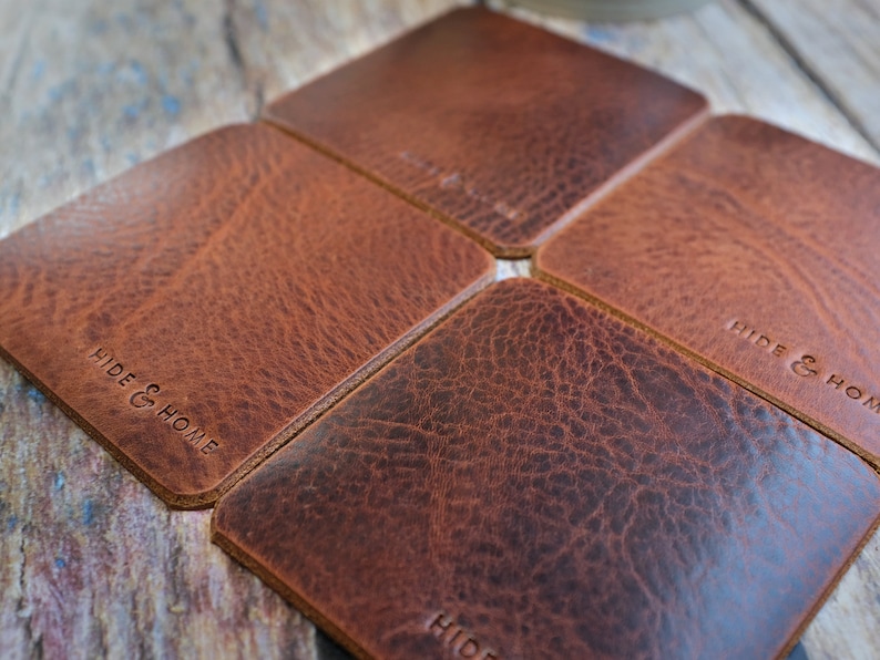 May include: Four square brown leather coasters arranged in a flower-like pattern. The coasters have a textured surface and the words "HIDE & HOME" are embossed on the surface. The coasters are a warm brown color.