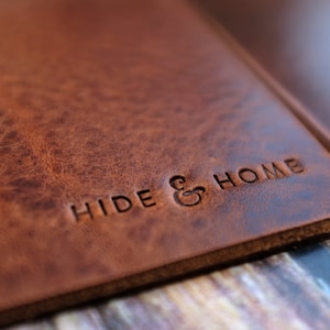 May include: Close-up of a brown leather product with the embossed text "HIDE & HOME". The leather has a textured, distressed appearance. The product appears to be a notebook cover or a similar item.