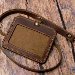 May include: A brown leather ID badge holder with a rectangular window and a matching leather neck lanyard. The holder features visible stitching along the edges, a brass-toned metal rivet at the top, and a swivel lobster clasp attachment.