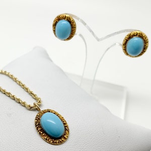 May include: A gold chain necklace with a blue oval gemstone pendant set in a gold filigree frame.  The necklace is displayed on a white surface.  Next to the necklace are a pair of gold stud earrings with blue oval gemstones set in gold filigree frames. The earrings are displayed on a clear stand.