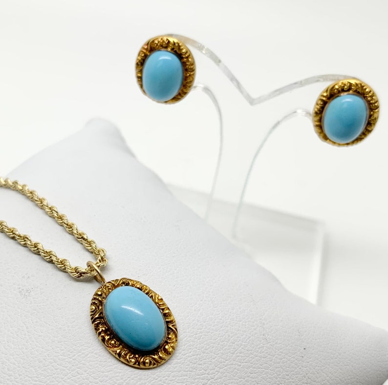 May include: A gold chain necklace with a blue oval gemstone pendant set in a gold filigree frame.  The necklace is displayed on a white surface.  Next to the necklace are a pair of gold stud earrings with blue oval gemstones set in gold filigree frames. The earrings are displayed on a clear stand.