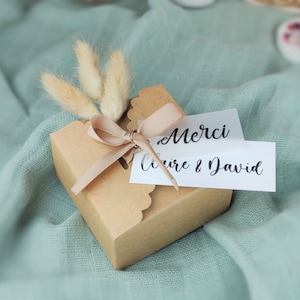 Peut inclure: Une petite boîte cadeau marron avec un bord festonné, nouée avec un ruban beige. La boîte est ornée de fleurs beiges séchées et duveteuses et d'une étiquette blanche avec l'inscription "Merci Claire & David" en écriture noire.