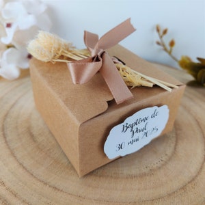 Peut inclure: Une petite boîte cadeau en carton brun, nouée d'un ruban marron clair et ornée de fleurs séchées. Une étiquette blanche avec le texte "Baptême de Paul 30 mai 2025" est apposée. La boîte est posée sur une surface en bois.