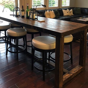 May include: A long, rustic wooden table with a black metal frame and four black metal bar stools with white cushioned seats. The table is set with two lanterns and three bottles of wine.