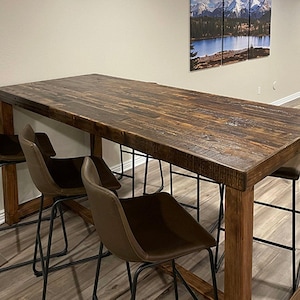 May include: A rectangular, dark brown wooden bar table with a distressed finish. Four brown leather bar stools with black metal legs are positioned around the table. The table is approximately 72 inches long. The room has light-colored walls and a wood-look floor.