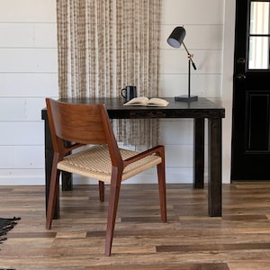 May include: A dark wood desk with a woven chair and a black lamp with a gold arm. The desk has a book and a mug on it. The desk is in front of a white wall with a woven wall hanging.