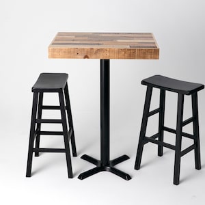 May include: A square, wooden-topped bar table with a black metal base, flanked by two black wooden bar stools. The table's top is made of reclaimed wood, and the stools have a simple, ladder-back design.