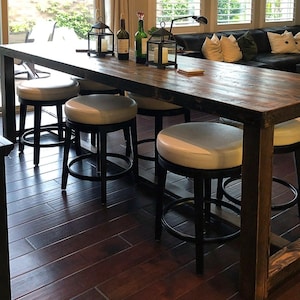May include: A long wooden table with a rustic finish and black metal legs. Four black metal bar stools with white padded seats surround the table. The table is set with candles and wine bottles.