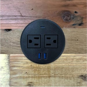 May include: A black circular power outlet with two standard electrical outlets, two USB ports, and a USB-C port. The outlet is set into a wooden surface with a natural wood grain pattern.