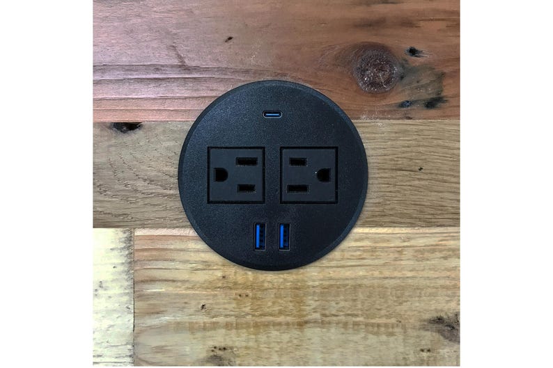 May include: A black circular power outlet with two standard electrical outlets, two USB ports, and a USB-C port. The outlet is set into a wooden surface with a natural wood grain pattern.