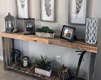 May include: A rustic wooden console table with a lower shelf. The table is decorated with a variety of items, including a framed photo, a potted plant, a lantern, and a decorative object. The table is set against a wall with three framed prints of feathers.