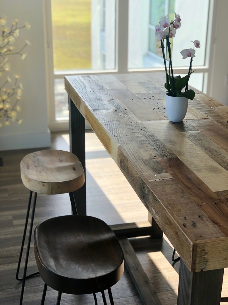 May include: A rustic wooden dining table with a natural finish, paired with two wooden stools with black metal hairpin legs. A white pot with an orchid plant sits on the table. The table is approximately 72 inches long.
