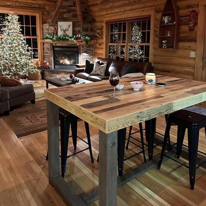 May include: A rustic wooden bar table with a light gray metal base, paired with black metal stools. The table is in a cozy cabin setting with a fireplace, Christmas tree, and leather sofa.