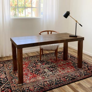 May include: A dark wood desk with a white chair and a black lamp with a gold arm. The desk is sitting on a red and black patterned rug.