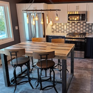 May include: A rustic wooden dining table with a black metal frame and four black metal bar stools. The table has a distressed wood finish and the stools have a black metal frame with a black leather seat. The table is set in a kitchen with white cabinets and a stainless steel stove.