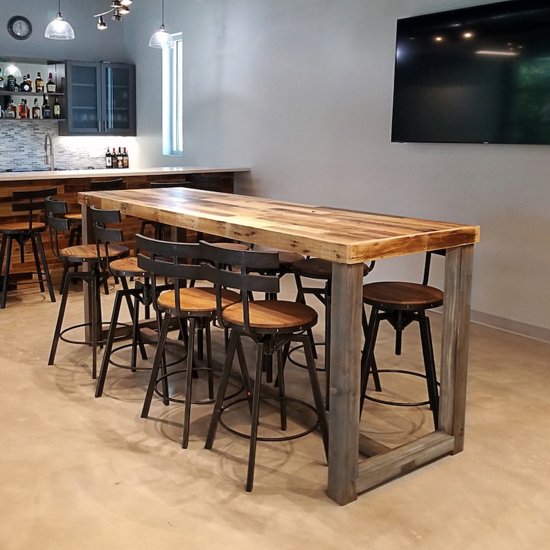 May include: A rustic wooden bar table with a light brown tabletop and gray legs. The table is surrounded by several metal and wood bar stools. The bar area also includes a bar with liquor bottles and a television.