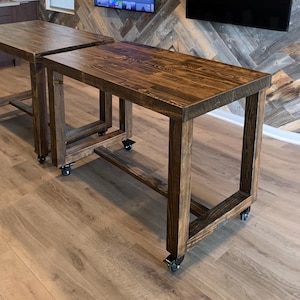 May include: Two wooden tables with a dark brown finish. The tables have a rectangular top and are supported by square legs. Each table is mounted on four caster wheels. The tables are placed on a light brown wooden floor.