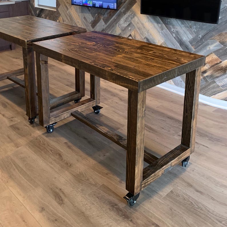 May include: Two wooden tables with a dark brown finish. The tables have a rectangular top and are supported by square legs. Each table is mounted on four caster wheels. The tables are placed on a light brown wooden floor.