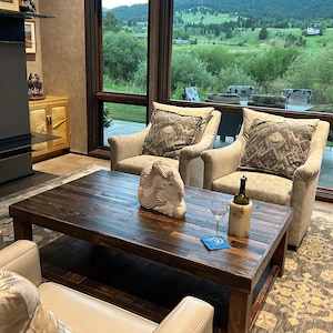 May include: A living room with a large window overlooking a green valley. Two beige armchairs with patterned cushions face a coffee table with a wooden sculpture and a bottle of wine. A beige rug covers the floor.