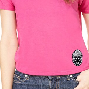 Könnte beinhalten: Ein pinkfarbenes T-Shirt mit kurzen Ärmeln und einem schwarz-weißen Buddha-Motiv auf der Vorderseite. Das Shirt wird von einer Person mit langen braunen Haaren und einer blauen Jeans getragen.