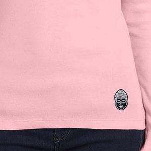 Puede incluir: Una camiseta rosa de manga larga con una pequeña cara de Buda bordada en la parte inferior derecha.