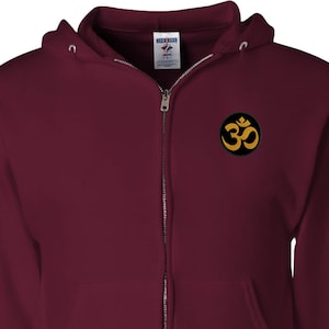 May include: A burgundy zip-up hoodie with a front pocket and a hood. The hoodie features a small, circular patch on the chest with a gold Om symbol on a black background. The zipper is silver.