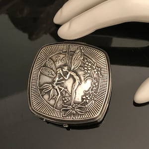 May include: A silver-toned, square compact with a detailed relief design. The design features a central image of two fairies surrounded by floral and leaf motifs. The compact has a hinged closure and a textured border.