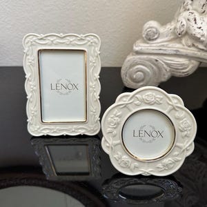 May include: Two off-white Lenox picture frames with raised floral and vine relief detailing. One frame is rectangular with rounded corners, and the other is circular. Both feature a thin gold-toned inner border surrounding the central display area.