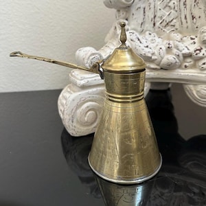 May include: A vintage brass coffee pot with a long handle and a conical body. The pot has a lid with a small finial and etched geometric patterns. The pot is likely used for making Turkish or Greek coffee.