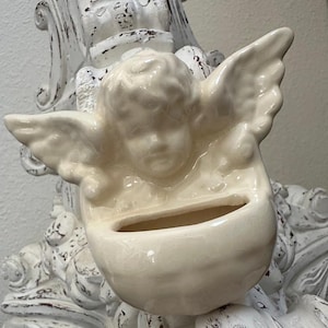 May include: Cream-colored ceramic angel-shaped planter with outstretched wings. The planter has a small opening for plants or flowers. The angel is set against a distressed white decorative stand.