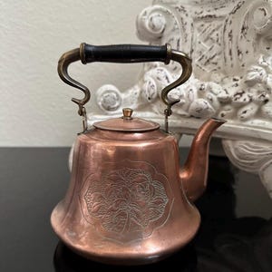 May include: A copper tea kettle featuring a wide, flared base and a curved spout. The body is adorned with an intricate, etched floral design centered on the front. It includes a brass handle with a black grip and a matching copper lid.