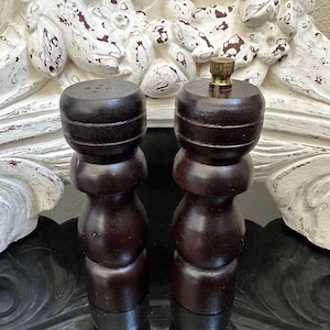 May include: A pair of dark brown wooden salt and pepper grinders with a cylindrical, tiered body design. One grinder features a small metallic knob at the top, while the other has a flat top with small holes for dispensing seasoning.