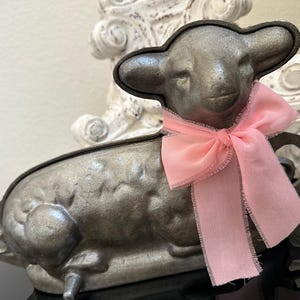 May include: A vintage metal lamb-shaped mold with a light pink ribbon tied around its neck. The lamb has a textured surface and is resting on a black surface. The mold is likely used for baking or decorative purposes.