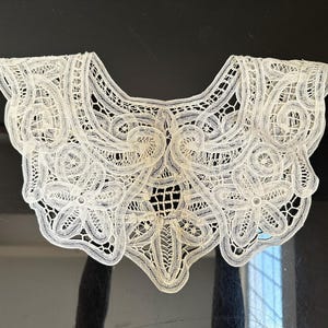 May include: An off-white, intricately designed lace collar. The collar features a scalloped edge and a detailed openwork pattern with floral and geometric motifs. The delicate lace is displayed against a dark background, highlighting its fine craftsmanship.