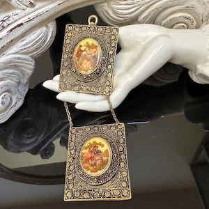 May include: A gold-toned metal double pendant necklace featuring two rectangular plates with intricate floral filigree borders. Each plate contains a central oval medallion depicting a colorful pastoral scene with figures in a landscape setting.